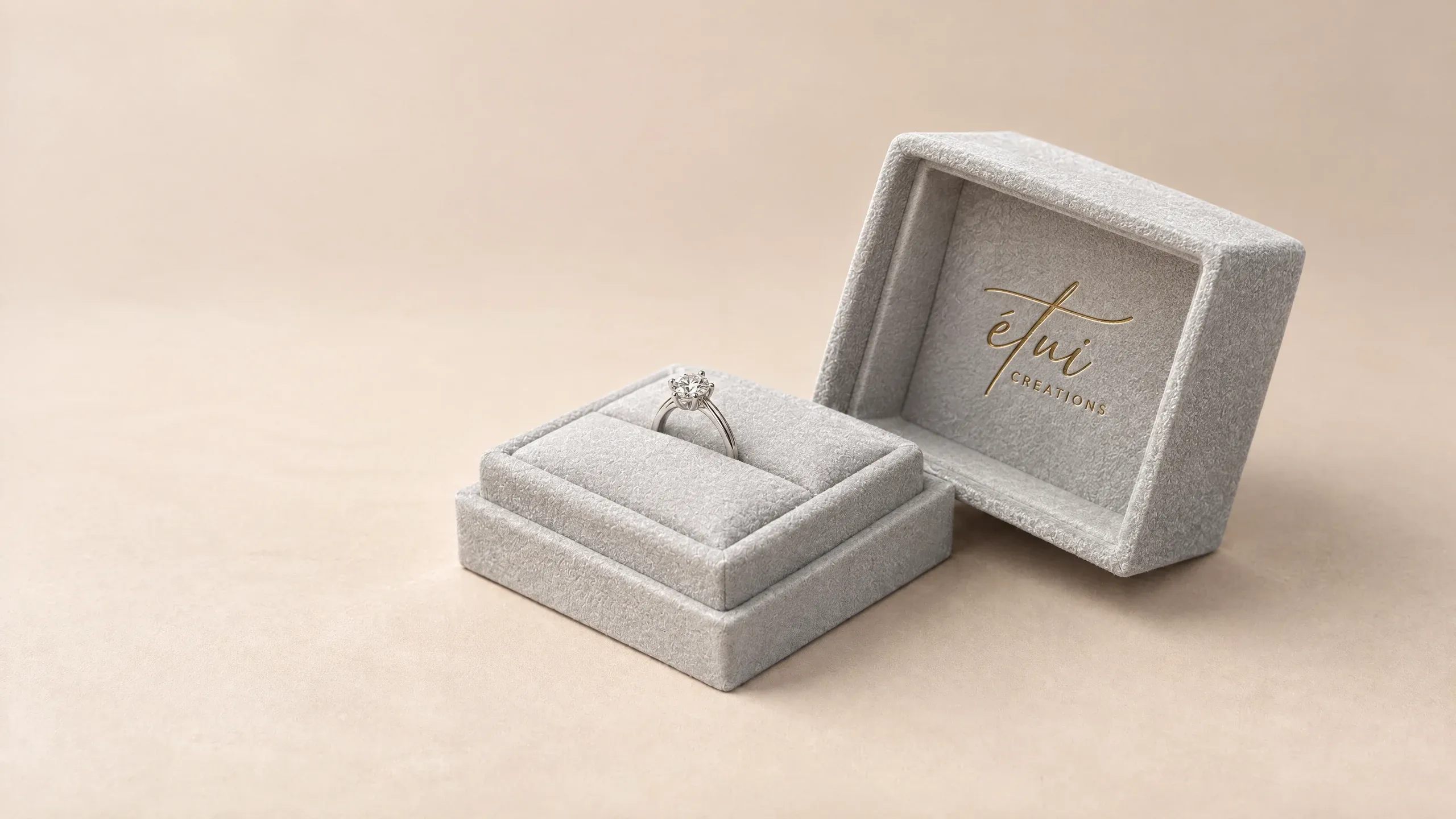 Open light-grey suede ring box with a diamond ring and the gold-stamped étui logo in the lifted-off lid